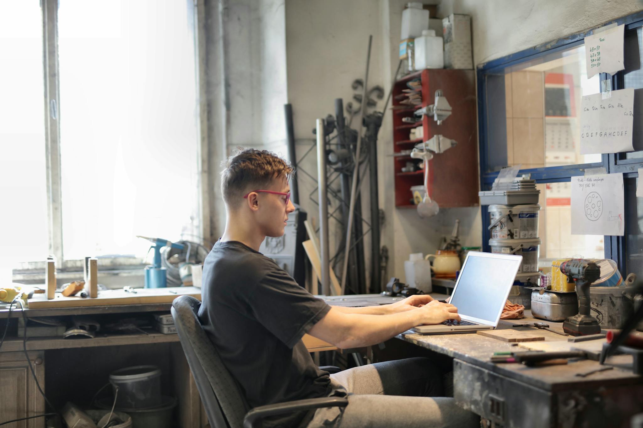 A young man works on a laptop in a creative workshop setting, surrounded by tools and materials.